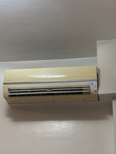 The image shows a close-up, eye-level view of a light beige Sharp air conditioning unit mounted on a light gray wall, likely in an indoor setting. The air conditioning unit is the primary subject, filling the majority of the frame. It has a horizontal vent at the bottom with adjustable louvers and a digital display panel on the right side showing a blue screen with what appears to be temperature settings and several indicator lights, some of which are illuminated red. The text "SHARP" is visible on the left side of the unit, and "ECO Inverter" is printed on the right. The wall has a slightly textured finish and is painted a neutral, light color. The ceiling is visible at the top of the image, also a light color. The lighting in the scene appears to be artificial, casting a soft glow on the surfaces. There are no people or animals present in the image. The overall scene suggests a typical residential or commercial interior space. Given the context of Pasay City, Philippines, this could be a room in a hotel, condominium, or office building.