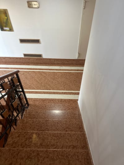 The image depicts a set of marble stairs with a brown and white speckled pattern. There are two white stripes running horizontally across the visible steps. A dark metal railing with wooden accents is visible on the left side of the stairs. The walls are white, and a framed picture is on the upper left wall, with two rectangular air vents below it. A door frame with hinges is visible on the upper right. The scene is indoors, likely in a building's common area or staircase, and is devoid of any people or activity. The location is not specified as San Martino Buon Albergo, Italy, in the image provided.
