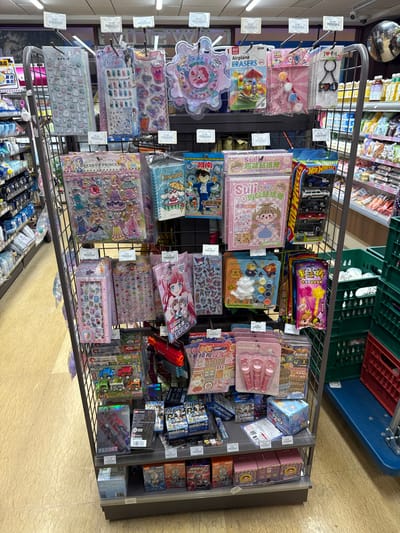 A vertical image captures a multi-tiered metal retail display rack within a convenience store or supermarket located in Tongxiang, China. The rack is densely stocked with various consumer goods, primarily children's stationery, small toys, and craft supplies, presented on hooks and shelves.

The top tier displays hanging items including numerous sticker sheets featuring diverse designs such as cartoon mushrooms and decorative motifs, alongside "My Little Pony" themed merchandise, "Airplane Erasers," small craft kits, and assorted keychains. Below, the middle tiers feature "Dress Up Princess" sticker books, "Detective Conan" branded notebooks, "Suli DIY" sticker books, a Hot Wheels five-pack of die-cast cars, additional sticker collections with anime-style characters, small paint and mold craft kits, toy wands, and a prominent orange toy blaster. The bottom shelves are stocked with various boxed items, including what appear to be trading card packs or blind boxes (some depicting "Ultraman"-style characters), small boxed figures, and sets of pink cosmetic applicators. Price tags are visible above many items, displaying specific values such as 9.90 or 19.90.

The store environment features overhead fluorescent lighting and a light-colored tiled floor with a subtle geometric pattern. In the background, fully stocked shelves of general merchandise, including packaged foods and beverages, are visible. To the right of the display stand, green and red plastic crates are stacked on a blue utility dolly. A convex security mirror is affixed in the upper right portion of the frame. No individuals are present in the immediate foreground or actively interacting with the display.