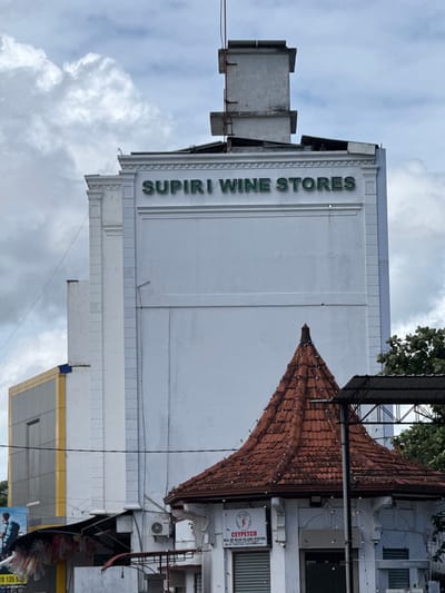 The image displays an urban street scene in Matara, Sri Lanka, dominated by several buildings under an overcast sky. The photograph is oriented vertically, showing the side of a prominent white, multi-story building on the left. This building features a large, green sign running vertically along its facade that reads "SUPIRI WINE STORES". The architecture of this building includes decorative molding and a flat roofline.
To the right of the "SUPIRI WINE STORES" building, a distinct building with a red-tiled, steeply pitched roof is visible. The roof has numerous small lights or decorations strung across its surface, suggesting it might be an entrance or a distinctive architectural feature. Above this roof, green foliage from a tree is visible.
Further to the right, behind and partially obscured by the tiled roof building, is another structure. On its facade, a white rectangular sign with text and a logo is clearly visible. The sign reads "CEYPETCO W.A. DE SILVA FILLING STATION" with an address and phone number below: "No. 129, Main Street, Matara. Tel. No. 041-2222247". The CEYPETCO logo, featuring a stylized elephant and gas pump, is also present. This indicates a petrol station. Various wires and utility lines crisscross the upper parts of the scene.
Below the "SUPIRI WINE STORES" building and extending to the bottom right of the frame, another building with yellow trim and gray panels can be seen, along with what appears to be a section of a billboard or advertising poster featuring a person. The overall impression is a bustling commercial area, likely taken during the daytime on a cloudy day.