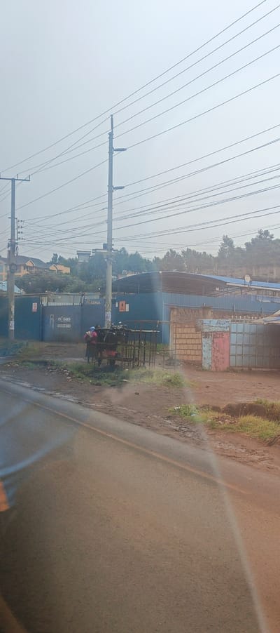 A street scene shows a gray road surface with a hint of rainbow. In the background, power lines stretch across a hazy sky. A blue building with "m Gas" lettering is visible. People stand by a metal cage-like structure. Faded brick and metal structures are next to the blue building. Greenery and bare dirt line the road's edge.