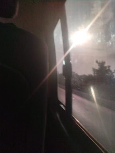 This is a blurry, low-light image taken from inside a vehicle, looking out through a window. The sun is visible in the upper right quadrant, creating lens flares and glare that obscure details. The foreground is dominated by the dark interior of the car, possibly a seat and a window frame. The background shows a road with trees and buildings on either side, suggesting an urban or suburban environment. The overall impression is that of a candid, possibly accidental, shot taken while in motion. The location in the city of Mayo, Nigeria, is not discernible from the visual information provided in the image.