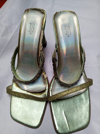 Two worn, green and gold high-heeled sandals sit on a white surface. The square-toed shoes have a strap across the front and a decorated strap over the top of the foot. The finish on the shoes is worn, with some areas peeling and discolored. Each insole displays a logo near the heel. The overall condition suggests they are used and somewhat aged.