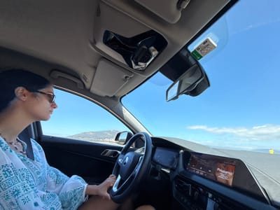 A female driver, seen in profile, operates a left-hand-drive vehicle, identifiable as a modern BMW by its steering wheel logo and dashboard design. She has dark hair, wears sunglasses, and a light-colored tunic with a teal geometric pattern. Her left hand is positioned on the steering wheel. The car's interior includes a digital instrument cluster and a central infotainment screen displaying "Positions" by Ariana Grande. A white electronic toll device, branded with green text, is affixed to the upper-right portion of the windshield. The exterior view, captured from the passenger side, shows a clear blue sky with scattered white clouds over a body of water. In the distance, a landmass with vegetated hills is visible, consistent with the coastal and riverine landscape of Caminha, Portugal, likely viewed from a road near the Minho River estuary or along the Atlantic coast approaching or within the municipality.