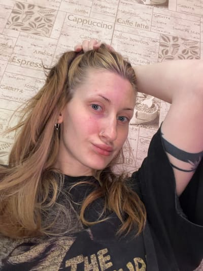 A young Caucasian woman with blonde hair and blue eyes is pictured in a selfie. She is wearing a black t-shirt. Her left hand is in her hair, and her right arm is bent behind her head, revealing a black tattoo on her upper arm. The background is a beige wallpaper with a pattern of coffee-related words and imagery, such as "Espresso," "Cappuccino," and coffee beans. The image appears to be taken indoors.