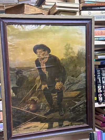 Here's a description of the image:
The image shows a framed painting, positioned amongst a collection of books. The painting is a dark and weathered depiction of a man in a hat, seated near a campfire. The man is wearing a dark coat and appears to be engaged in some activity near the fire. Around him, there are implements suggesting a camp scene, like a pot or jug. The surrounding environment is indistinct, hinting at a rural or outdoor setting. The painting is housed in a dark brown frame with a gold inner lining. The books around the painting are various colors and sizes. The location of the image is in the city of Nikolo-Beryozovka, Russia.