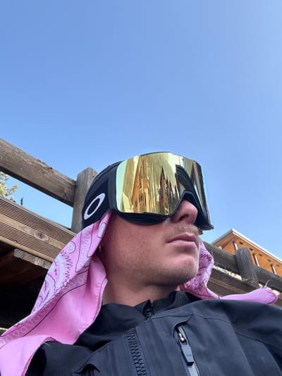 A male individual is depicted from the mid-chest upwards, looking to the right of the frame, under a clear blue sky in Saint-Chaffrey, France. He wears black Oakley brand ski goggles with a wide, reflective gold-mirrored lens over a black beanie. A light pink bandana with a white paisley pattern is positioned beneath the beanie, with ends visible on both sides of his neck/jawline. The individual has a light mustache and stubble, and a silver hoop earring is visible in his left ear. He is wearing a black jacket with a prominent vertical zipper on the chest and a smaller horizontal zippered pocket on the upper left chest.

In the background, to the left, a wooden railing structure composed of horizontal planks and vertical posts is visible, with sparse green foliage behind it. To the right, the corner of a building with wooden siding and a sloped roofline can be seen. The reflective goggles prominently display a distorted reflection of a large, multi-story structure with distinct alpine-style architecture, characterized by multiple gabled roofs and visible windows, consistent with buildings found in a mountain resort environment such as Saint-Chaffrey.