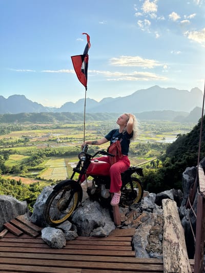 A woman with blonde hair is sitting on a red motorcycle. She is wearing a black t-shirt, red pants, and pink slides. Her left hand is on her head, and she is looking to the left. The motorcycle is on a rocky surface with a wooden platform to the right. A flag is attached to a pole on the left side of the motorcycle and is waving in the wind.

The background shows a vast landscape of green rice fields and mountains under a bright blue sky with scattered clouds. The scene appears to be at a high vantage point, possibly a viewpoint or a cliff. The lighting suggests it is daytime, likely late afternoon due to the warm glow on the clouds and mountains. The overall mood is adventurous and scenic, with a sense of freedom and exploration. The flag appears to be the flag of Laos. There is no visible text in the image.