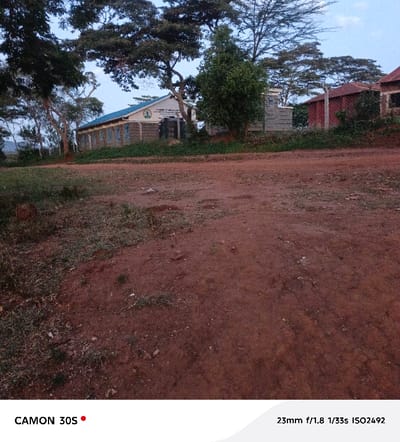 A dusty, reddish-brown ground dominates the foreground. In the middle ground, a one-story building with a blue roof stands flanked by trees. A wall separates the building from a red-roofed structure further to the right. The sky is light blue and overcast. The image is taken with a CAMON 30S at 23mm f/1.8 1/33s ISO2492.