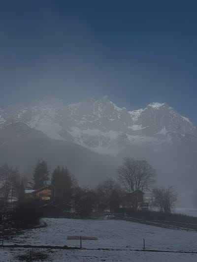 This image captures a snow-covered winter scene in Going am Wilden Kaiser, Austria. The upper third of the frame features a clear, deep blue sky. Below, the rugged, snow-capped peaks of the Wilder Kaiser mountain range dominate the background, partially obscured by a pervasive atmospheric haze that extends into the midground. In the midground, a cluster of buildings, typical of alpine architecture, is visible, interspersed with bare deciduous trees and dark evergreen conifers. One timber-clad building displays a warm yellow illumination from its windows. The foreground consists of a flat, snow-covered field, traversed by faint fences and featuring a horizontal wooden plank-like object resting on the snow. No individuals or overt activities are present. The overall scene suggests a cold, possibly misty, winter day.