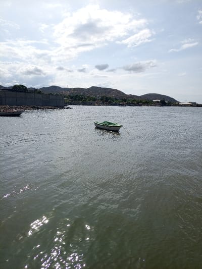 A small, green and white rowboat floats in a tranquil body of water, tethered by a rope. Sunlight glints off the water's surface, creating shimmering reflections. A concrete barrier lined with dark stones borders the water's edge on the left. Rolling, green hills and a hazy sky form the backdrop, with a hint of buildings visible along the distant shore. The sky is partly cloudy.