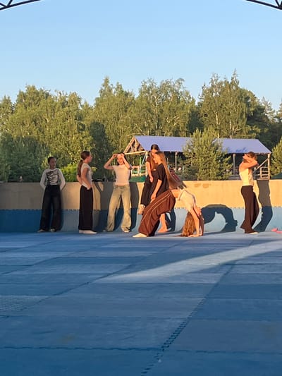 This image captures a group of people, likely teenagers, engaged in a physical activity on a blue matting surface. The subjects are positioned in a line, performing what appears to be a synchronized dance or gymnastics routine, characterized by arched backs and bending poses. The scene is outdoors, with a clear blue sky overhead and lush green trees visible in the background. The lighting suggests it is daytime, likely late afternoon or early evening, given the long shadows cast by the individuals.
The individuals are dressed in casual athletic wear. One person wears a white t-shirt and dark pants, another a white t-shirt and jeans, and a third person is in a black top and brown pants. Further down the line, other participants are visible in similar attire. The blue matting has a grid-like pattern, indicating it's a designated area for activities like sports or performances. A structure, possibly a small stage or seating area, is partially visible behind the trees. The overall atmosphere appears to be focused and collaborative, with the participants concentrating on their movements. The location context provided is Krasnoturansk, Russia. There is no visible text in the image.