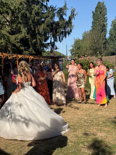 The image depicts a wedding party gathered outdoors in what appears to be a garden setting. The bride, in a voluminous white gown, stands facing away from the camera, her back adorned with intricate white floral details. She holds a bouquet of colorful flowers. Surrounding her are several women, presumably bridesmaids and guests, dressed in various colored gowns and dresses. The attire ranges from a shimmering gold mini-dress and a long, flowing orange dress to pastel pink and yellow floor-length gowns, and a bright, multi-colored pink, orange, and yellow ombre dress.
The environment is characterized by lush green trees providing shade, a clear blue sky, and dry, sun-drenched grass. In the background, a wooden pergola decorated with string lights suggests a venue for celebration or reception. There are also rose bushes visible in the middle ground. The lighting indicates it is daytime, likely late afternoon due to the long shadows.
While the image is rich in visual detail, there is no specific information within the image itself to pinpoint the location to Hem, France. The scene is a generic outdoor wedding celebration.