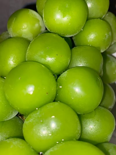 This is a close-up, high-angle photograph of a cluster of bright green grapes. The grapes are plump and round, with a glossy sheen that suggests they have been recently washed or are naturally dewy. Small water droplets are visible on the surface of many of the grapes, catching the light and adding to their fresh appearance. The lighting is bright, highlighting the vibrant green color and the smooth texture of the fruit. Some of the grapes have faint lines or subtle imperfections, adding to their natural look. The background is dark and out of focus, ensuring that the grapes are the sole subject of the image. No people, text, or specific setting details are visible, other than the grapes themselves. The image appears to be taken indoors or in a shaded area, given the artificial-looking lighting and lack of harsh shadows. The focus is entirely on the texture, color, and spherical shape of the individual grapes and their collective arrangement.