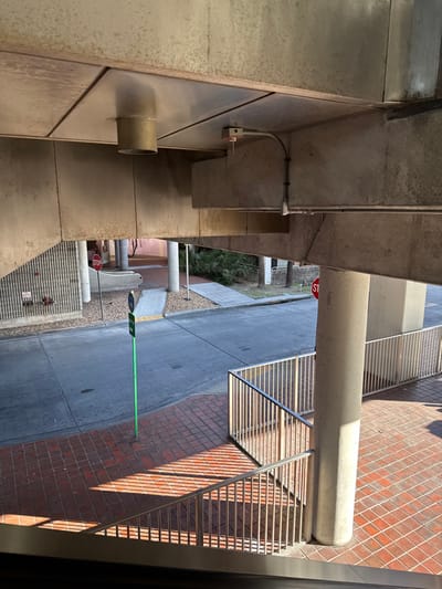 The image presents an elevated, downward-angled view of an outdoor public area located under an overhead concrete structure, identified as part of the Hialeah Market Station or Hialeah Station of the Miami-Dade Metrorail system in Hialeah, Florida, United States of America.

The foreground displays a reddish-brown square paver or brick surface, with strong parallel shadows cast across it by silver-colored metal railings that form a barrier or stairway descending out of frame to the lower left.

The mid-ground features a grey asphalt or concrete roadway. To the left of the roadway, a light grey concrete sidewalk transitions into a ramp. Adjacent to this, a vertical wall with a ribbed texture, possibly concrete or metal, features a red fire hydrant. A red hexagonal STOP sign on a dark pole is positioned near the left sidewalk. A light green pole, topped with a small dark blue and white sign, stands at the edge of the roadway. On the right side of the mid-ground, a second light grey concrete path or ramp is visible, bordered by sparse green vegetation including palm trees. A second red hexagonal STOP sign is partially visible behind a large, round concrete support column on the right.

The overhead structure consists of exposed, unpainted concrete beams and slabs. The underside of the ceiling includes metallic panels. A cylindrical, light-colored metal vent or pipe extends downwards from the ceiling. A red-tipped fire sprinkler head or sensor is mounted on a concrete beam, connected by a curved metal conduit. Additional metal conduits run along the concrete support beams. Multiple large, round concrete columns support the elevated structure, one prominently on the right side of the frame.

No individuals, vehicles, or animals are present within the depicted scene.