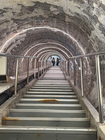 The image shows an interior perspective of a tunnel in Boj-Cătun, Romania, featuring a walkway flanked by railings. The tunnel's walls are textured with a gray, rocky material that appears to be naturally formed or hewn, with white, crystalline deposits along the surface. Illuminated rings follow the curve of the tunnel. The walkway is composed of gray panels or slabs. Two individuals are visible at the far end of the tunnel. The railings are constructed of silver-colored metal bars. Pipes or conduits run along the base of the walkway. The lighting within the tunnel is a combination of the illuminated rings and natural light filtering through the tunnel.