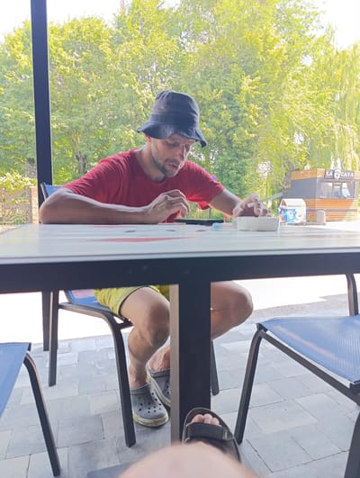 A man wearing a red t-shirt, a bucket hat, and yellow shorts sits at a table. He is smoking a cigarette and appears to be looking down at the table. In front of him is an ashtray and a few small objects. He is sitting on a blue chair. The table has a white surface with some colorful markings.
The setting appears to be outdoors, possibly a cafe or restaurant patio, with trees in the background. There is a building with the text "LA CAVA" visible behind the man. The weather seems to be sunny, as indicated by the bright lighting and the lush green foliage. The time of day appears to be daytime. The overall atmosphere seems casual and relaxed.