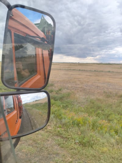 The image, taken from the perspective of a vehicle's side mirror, captures a view of a rural landscape in Budarinsky rural district, Kazakhstan. The foreground is dominated by the orange side of a truck, with its side mirror reflecting the scene. The main mirror, positioned at the top, shows a partial view of the truck's cabin and a glimpse of machinery behind it. Below, a second, larger mirror reflects the road and the surrounding dry, grassy fields stretching towards a distant horizon under a cloudy sky. The sky itself is filled with heavy, grey clouds. The road appears to be paved, with sparse green and yellow vegetation lining its edges. The overall impression is one of a journey through an open, agricultural area. There are no visible people in the scene.