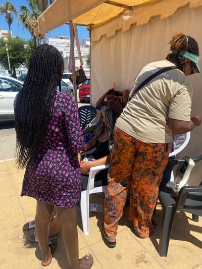 An exterior shot, taken on a sunny day in Conil de la Frontera, Spain, depicts three individuals under a pale yellow awning. The foreground is dominated by a young woman from the rear, sporting long, dark braids and wearing a black dress with a pink floral pattern. Beside her, and slightly behind, is another person seated in a white plastic chair, their face obscured, wearing a black top and dark pants. To the right, a third individual stands, facing away from the camera, dressed in a light tan t-shirt with text on the back and brightly patterned orange and green pants. This person appears to be attending to the seated individual, perhaps braiding their hair. The ground is paved with yellow tiles, and sunlight casts shadows. In the background, a white car and a red car are visible, along with buildings and palm trees. A Louis Vuitton bag hangs from the awning.