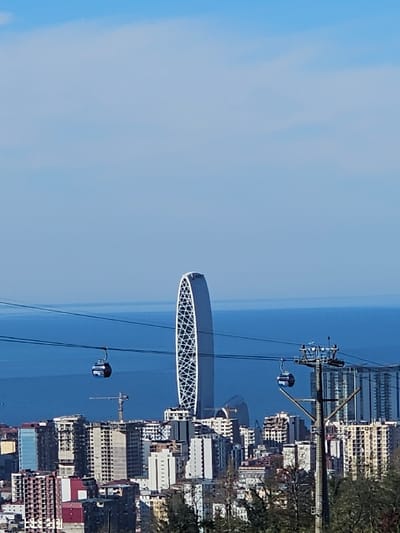 The image presents a panoramic daytime view of Batumi, Georgia, from an elevated perspective, overlooking a dense urban area towards the Black Sea. A prominent, tall, oval-shaped skyscraper, the Batumi Tower, with a white, lattice-patterned facade, stands centrally in the mid-ground. An aerial cable car system traverses the scene horizontally, featuring dark cables from which two blue gondolas are suspended. One gondola is positioned to the left of the Batumi Tower, while another is to the right, near a grey support pylon. The cityscape below the cable car lines consists of numerous multi-story buildings, displaying diverse architectural styles, with evidence of ongoing construction including a yellow tower crane. The deep blue expanse of the Black Sea forms the horizon line behind the city, meeting a clear, light blue sky with minimal cloud cover. The immediate foreground shows the upper parts of dark green foliage.
