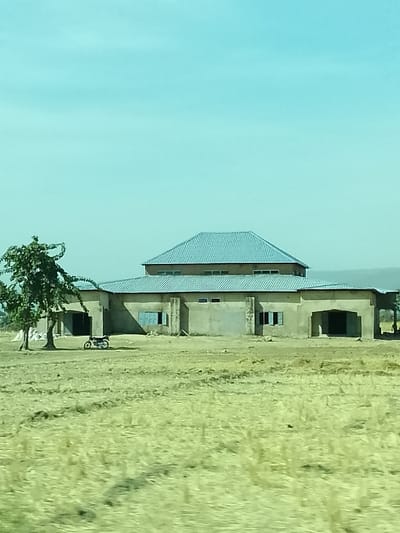 In Minting Chip, Nigeria, the image depicts a daytime scene featuring a multi-sectioned building situated in a dry, open field under a clear, light blue sky. The central building consists of a lower, single-story concrete structure with an unpainted, light grey finish, appearing to be in an unfinished state. This lower section has multiple rectangular window openings, some featuring light blue frames or shutters. A large, arched entrance is visible on the right side, and another opening is partially obscured by a tree on the left. Rising above the center of the lower structure is a taller, second story with a light blue corrugated metal hip roof and a series of smaller rectangular windows below the roofline. To the left of the building stands a green, leafy deciduous tree with a visible trunk and branches. Parked on the dry, yellowish-green ground near the tree is a dark-colored motorcycle. The foreground consists of sparse, dry vegetation, resembling a harvested or fallow field. The background shows a faint horizon with distant, hazy landforms on the right. No people are clearly visible, and no discernible actions or interactions are occurring.