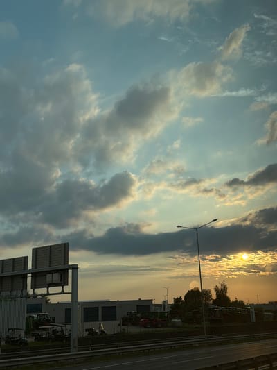 This is a landscape photograph taken at sunset in Kuří, Czechia, depicting a view from the side of a road. The sky is filled with a mix of light and dark clouds, with the setting sun casting a golden glow. In the foreground, a highway with a metal guardrail is visible. Beyond the road, a lot is filled with various agricultural vehicles, including tractors and smaller utility vehicles. A tall lamppost stands in the middle ground, and buildings with large garage doors are situated behind the vehicles. Trees are scattered throughout the scene, and some advertising signs are also present. The overall impression is of an industrial or agricultural area during a beautiful sunset.