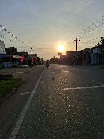 An urban scene in Beunot, Indonesia, features a street extending into the distance towards a setting sun. Power lines are strung above the buildings on both sides of the street. Red banners and storefronts are visible along the left side. A lone figure on a motorbike is centered on the road. The asphalt road is marked by white lines. Vegetation grows along the road's edge.