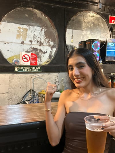 A young woman with long dark hair, wearing a strapless brown top and jewelry, holds a partially filled clear plastic cup containing a yellow liquid (likely beer) and smiles at the camera. She is positioned behind a bar, which is made of light-colored wood.
The background features a wall with exposed concrete and wiring, typical of a casual bar environment. Mounted on the wall, there's a black metal structure with two round, reflective "porthole"-like openings. A round sticker with a no-smoking symbol and the text "PROHIBIDO FUMAR, ESPACIO 100% LIBRE DE HUMO DE TABACO, EN CASO DE INCUMPLIMIENTO DE LA LEY EN EL ESTABLECIMIENTO, DENUNCIA 800 033 5050 SALUD COFEPRIS" is partially attached to one of the openings. A vertical green sign is adjacent to the round metal feature with the text "MEXICO 15 EL QUELITE SOY VIRGEN TE LO JURO POR MIS HIJOS."
Based on the sign "SOY VIRGEN TE LO JURO POR MIS HIJOS," the location is likely in the Quelite area near Mazatlán, Mexico, where such signs are commonly seen in local establishments.
