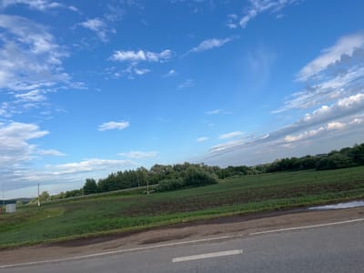The image shows a landscape scene in the city of Zainsk, Russia. In the foreground, a paved road runs horizontally across the bottom of the frame. To the left of the road, a grassy embankment slopes upwards, with some dark soil patches visible. Beyond the embankment, a vast green field stretches out, appearing to be agricultural land. In the middle ground, a dense line of various trees forms a natural border. Further in the background, rolling hills are visible under a bright blue sky dotted with fluffy white clouds. A small utility box is located on the left side of the embankment, and streetlights can be seen along the tree line. The scene is devoid of any people or animals. The overall impression is that of a rural or semi-rural area in Zainsk.