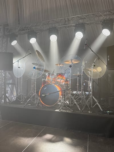 The image depicts a drum set on a stage bathed in bright light. The set features multiple drums, cymbals, and clear drum shields. Several stage lights are visible on the left, casting strong beams. The location is likely indoors, as suggested by the stage setup and lighting. There is no reliable evidence in the image to confirm that the scene is in the city of Shchekutino, Russia.