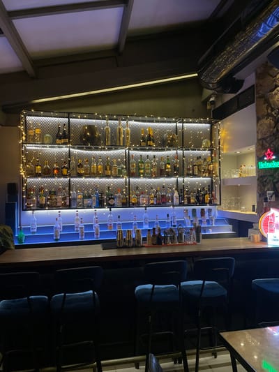 This is an interior photograph of a bar or pub in Chaidari, Greece, taken from a slightly elevated perspective. The image is oriented vertically (90 degrees rotated from normal viewing).
The central focus is the well-stocked bar area. On the left side, a large shelving unit is filled with numerous bottles of spirits and wines, arranged in individual compartments. This unit is adorned with warm white fairy lights, creating a sparkling, illuminated display. To the right of this, another section of the bar features a clean, modern display of clear bottles, likely spirits, illuminated from below by cool blue LED lights, setting them apart. Several silver cocktail shakers and other bar tools are visible on the counter.
To the far right, a dark bar counter is lined with multiple bar stools featuring cushioned blue seats. This seating area appears darker, contrasting with the brightly lit bar displays.
In the upper background, a rugged stone wall is visible. Above the bar area, a green neon sign clearly displays the word "Heineken." Another round, illuminated sign with red and white colors is partially visible next to it, though any text on it is indistinct. A large, dark, industrial-style air duct runs across the top of the ceiling area.
No people are present in the image. The lighting suggests it is evening or nighttime, characteristic of a bar setting. The combination of warm fairy lights and cool blue LEDs, along with the rustic stone wall and modern bar elements, creates a dynamic and inviting atmosphere.