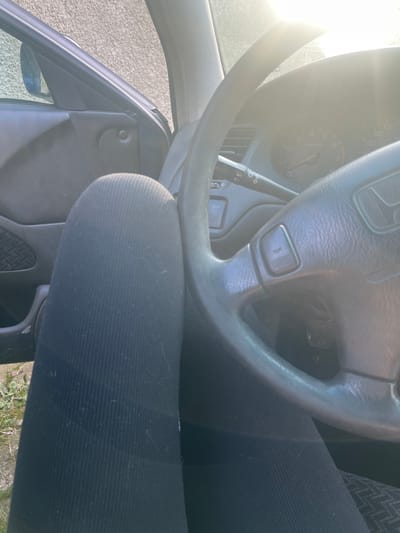 The image shows the interior of a car, focusing on the steering wheel, dashboard, and driver's seat. The steering wheel is a dark gray color with a Honda logo. The dashboard has a speedometer and tachometer. The driver's seat is black. The exterior is visible through the open car door. The location, Blaskovo, Bulgaria, cannot be determined from the image.