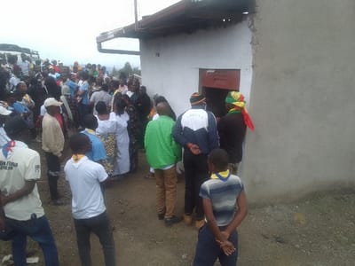A large crowd gathers near a building with a visible door. Individuals wear an assortment of clothing, including scout uniforms, traditional wraps, and casual attire. One person sports a colorful head covering and scarf. The crowd extends into the distance, suggesting a significant number of people present. The location appears to be on a slight incline with overcast weather.