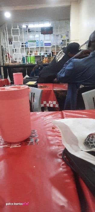The image portrays an indoor scene within an establishment in Homa Bay, Kenya, indicative of a casual eatery or a retail space offering refreshments. In the immediate foreground, a large, salmon-pink insulated mug with a lid stands on a red, glossy table surface that extends across the lower portion of the frame. Adjacent to the mug on the right, a crumpled white napkin rests on a black object, possibly a bag or electronic device.
The mid-ground reveals several individuals seated at tables. One person, positioned with their back to the viewer, wears a black t-shirt with a white graphic and leans forward over a table. To their immediate right, another individual is partially visible, dressed in dark blue attire. Further back, a third person in a dark blue shirt is seated at a table, facing towards the right side of the frame. These tables are covered with red tablecloths featuring a repeating white checkered pattern. Grey chairs are visible around the tables.
The background features a white modular shelving unit or room divider with square compartments. This unit is stocked with various items, including multiple large blue water dispenser bottles, smaller green and clear bottles, and other retail products. A red illuminated sign, possibly displaying text such as "OPEN NOW," is visible above the shelving. The ceiling is equipped with fluorescent lighting fixtures, and a ceiling fan is mounted centrally. The environment appears to be an active, artificially lit commercial interior.