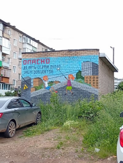 A mural is painted on the side of a brick building in Nytva, Russia, depicting two cartoon children taking selfies near power lines. The mural is rendered in a bright, simplistic style, with the words "ОПАСНО" (DANGER) in red capital letters at the top. Below this, in smaller text, it reads "ДЕЛАТЬ СЕЛФИ ОКОЛО ЭНЕРГООБЪЕКТОВ" (TAKING SELFIES NEAR POWER FACILITIES). The building itself is a light-colored brick structure, possibly a utility building or garage, with a metal roof overhang. To the left of this building is a multi-story apartment complex constructed of grey and beige brick. In the foreground, a dark grey sedan is parked on a gravel surface, partially obscured by tall green grass and vegetation. The sky is overcast. The mural serves as a public safety warning against dangerous selfie practices.