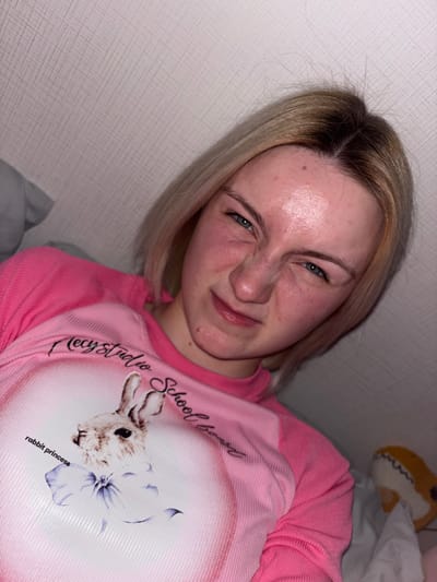 The image features a young woman, likely in her late teens or early twenties, captured indoors in Ligolambi, Russia. She has blonde hair with noticeable dark roots and blue eyes. Her skin displays some redness and minor blemishes. The woman is making a distinct scrunching or playful facial expression, narrowing her eyes and wrinkling her nose.
She is dressed in a pink long-sleeved shirt, which has a large, lighter pink oval-shaped graphic on the chest. This graphic features the head of a rabbit alongside a purple flower. Text visible on the graphic includes "Mecy Studio School" (partially obscured) and "rabbit princess."
The background suggests an indoor residential setting, possibly a bedroom, characterized by a light-colored, textured wall or headboard. In the upper right corner of the frame, a portion of a light-colored stuffed animal, which appears to be a shark with an open mouth and teeth, is visible. The lighting is artificial, illuminating her face softly. The photograph is taken from a slightly angled perspective, suggesting the woman is lying down or reclining. Her expression conveys a lighthearted and quirky mood.