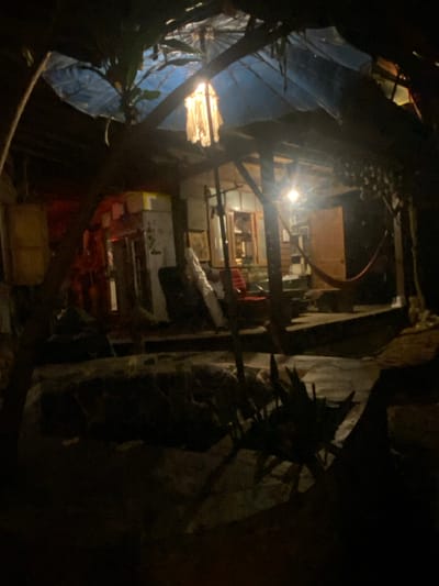 View from the exterior into the cluttered interior of a building in Baan Don Sai, Thailand. The foreground is dark, with what appear to be tree branches and foliage obscuring parts of the view. Through gaps, a cluttered interior is visible, containing furniture, boxes, and various other objects. Light emanates from within the building, illuminating some of the interior. The interior space appears to be multi-level. Overall, the scene has a chaotic and somewhat disorganized appearance.