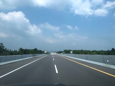 The image depicts a straight highway in Stabat, Indonesia, with a clear blue sky and scattered white clouds above. The road has multiple lanes, indicated by white and yellow lines. On either side of the highway, there are concrete barriers, beyond which are rows of palm trees and lush green vegetation. In the distance, power transmission towers are visible. The perspective is from a vehicle traveling on the highway, facing forward. There are no people or animals visible in the image. The overall environment suggests a rural or semi-rural setting on a highway.