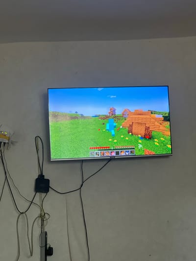 The image depicts a television screen displaying a scene from the video game Minecraft, located in a room in Şamaldı-Say, Kyrgyzstan. The game screen shows a pixelated landscape with a wooden structure, a light blue character, and red and yellow flowers. In the foreground of the room, a white router with several wires connected to it is mounted on the wall. Other cables are neatly bundled and run down the wall towards the floor, where they connect to black devices. The wall itself appears to be a light, textured color.