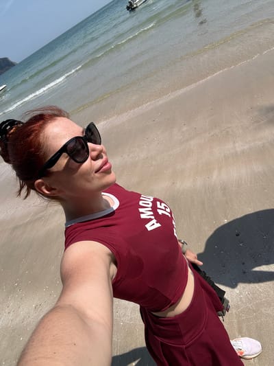 A woman is angled in the frame with her head at the bottom and her feet at the top. She wears a maroon top with white lettering "Amour 15", matching maroon pants, and white sneakers. She sports black sunglasses and has auburn hair pulled back in a bun. She appears to be standing on a sandy beach, with the ocean visible in the background on the left. A shadow is cast in the top right corner.