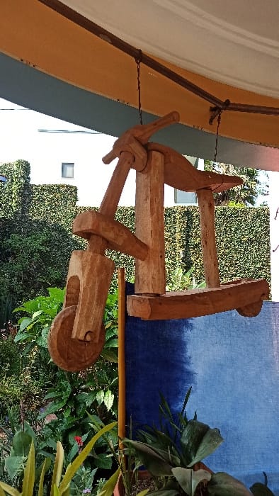 A light brown wooden object hangs by a thin wire. It has a wheel shape and a handle attached to it. In the background, there is a wall with a white window and dense green bushes. The objects are positioned in front of a navy blue fabric backdrop, lush green plants, and colorful flowers. The image is taken from a low angle.