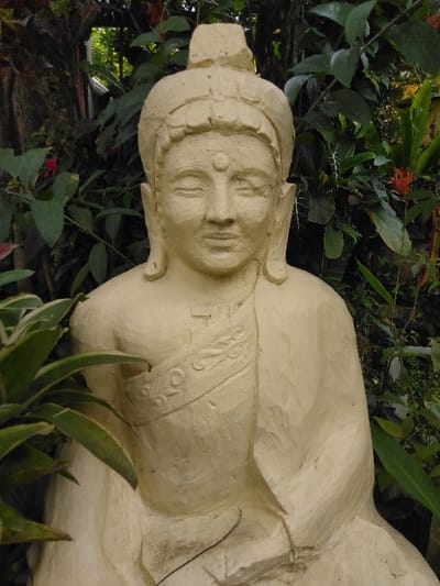 A light-colored, weathered statue, possibly carved from concrete or stone, is depicted centered in an outdoor setting in Ba Nwet Kone, Myanmar. The statue represents a seated figure with a serene expression, closed eyes, and an urna on the forehead, adorned with a distinctive headdress featuring a rectangular top. A robe drapes over the figure's left shoulder, leaving the right shoulder bare, and includes an embossed decorative band across the chest. The statue's surface exhibits a rough, textured finish with minor discoloration. The background is filled with dense, vibrant green foliage of various plants and trees, with some small red flowers visible among the greenery, and a broad-leafed plant partially obscures the lower left of the statue.