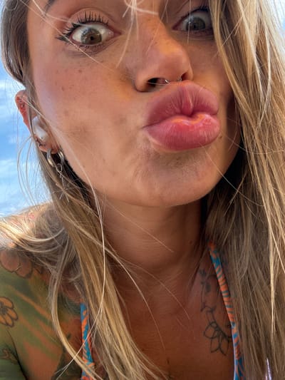 A close-up, low-angle, outdoor, full-frame shot shows the face of a blonde woman with a nose ring and ear piercings. She is puckering her lips towards the camera. Her eyes are wide open, looking directly at the camera. She is wearing a bikini top with blue and orange stripes. Her right shoulder and chest are visible, showing a colorful tattoo of flowers and other designs in green, orange, and yellow. Her blonde hair is blowing in the wind. The background consists of a bright blue sky. The scene appears to be the beach or poolside in Choeng Thale, Thailand.