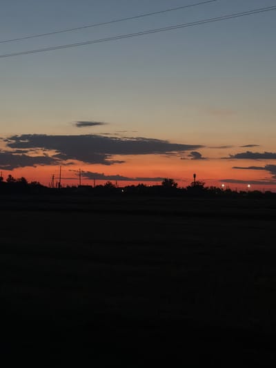 This is a photograph of a sunset over a rural landscape, possibly in or near Parkany, Moldova. The sky is a gradient of colors, transitioning from a light blue at the top to vibrant orange and red hues near the horizon. Several dark clouds are scattered across the sky. In the mid-ground, a silhouetted treeline is visible, interspersed with utility poles and distant lights. The foreground is almost entirely black, obscuring any details of the ground or vegetation. Two thin, dark lines, likely power lines, stretch horizontally across the upper portion of the sky. The overall impression is one of a peaceful, twilight scene in a countryside setting.