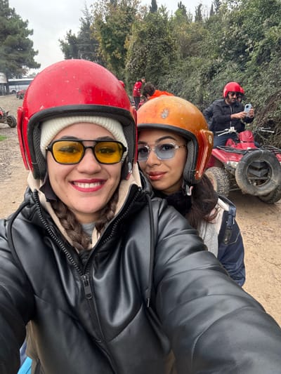 The scene depicts an outdoor gathering, likely for an ATV activity, located in Çiftlikköy, Turkey. In the foreground, two women are positioned for a selfie. The woman on the left wears a red helmet over a white beanie, yellow-tinted glasses, and a black zippered puffer jacket, exhibiting a wide smile with red lipstick. The woman behind her, to the right, wears an orange helmet, light blue-tinted glasses, and partially visible dark outerwear, also smiling with reddish-brown lipstick. In the midground, a man wearing a red helmet and dark jacket sits on a red ATV, holding a light-colored smartphone to take a photograph. Other individuals and red ATVs are visible further back on a dirt path. The background consists of dense green foliage, trees, and an overcast sky, with a white vehicle partially visible on the far left.