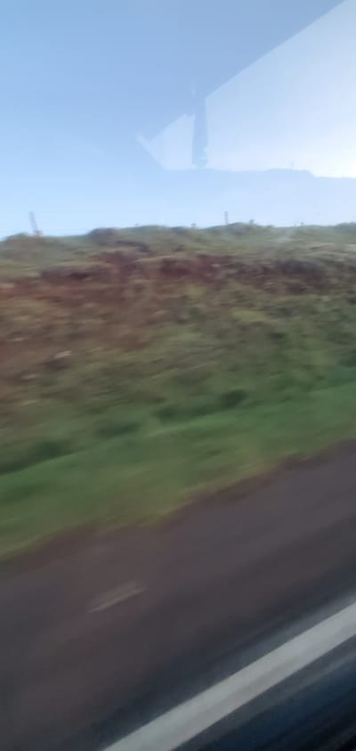 The image shows a view from a moving vehicle in what appears to be the Kisima ward of Kenya. The sky is visible on the left side of the image, with the shadow of an aircraft. A strip of land covered in brown and green vegetation runs down the center. A gray asphalt road is on the right side of the image. The overall picture suggests a rural environment along a road near the city.