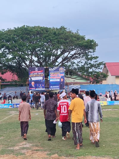In Desa Meunasah Blang, Indonesia, a group of young males, dressed in traditional attire, walk across a grassy field. Some wear sarongs and peci hats, others t-shirts and sarongs, with one young man in a red jersey bearing the name "MUZAMMIL" and the number "10." In the background, two large banners hang from trees, displaying text in Indonesian, and two buildings with red roofs are visible. A crowd of spectators is seated in tiered seating to the left, while others stand or mill about in the distance. The sky is overcast, and the overall atmosphere suggests an outdoor gathering or event.
