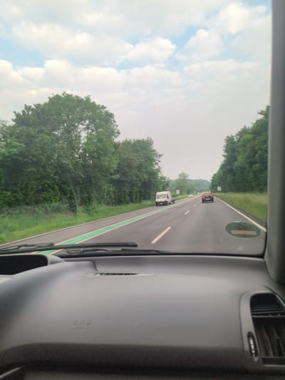 The image shows a view from inside a car traveling on a highway. The road has two lanes, with a green stripe along the left edge of the lanes. On the right is an orange car, followed by a white van pulling a trailer. There are trees on both sides of the road, and the sky is overcast. Based on the presence of green striped lanes, this may be a bicycle path next to the highway. Due to the available information, I can not confirm the exact location in the city of Borghorst, Germany.
