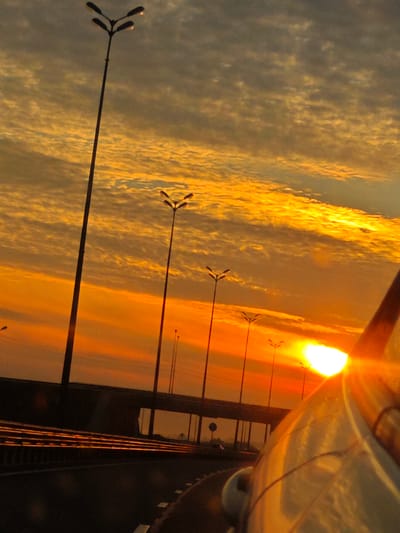 A view from a vehicle traveling on a highway during sunset. The sky is filled with a vibrant orange and yellow glow, with scattered clouds creating a textured pattern. Several tall streetlights, each with three lamps, are visible along the side of the road. The highway infrastructure, including a bridge or overpass, is dimly lit in the background. The right side of the frame shows the curved, reflective surface of the vehicle's body. The image captures a dynamic and colorful scene of a road at dusk.