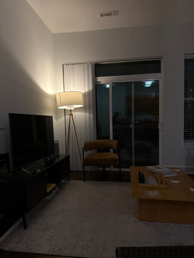 A living room scene is captured in a low-light setting. A large window with blinds provides a view of the outdoors. A tan chair sits near a coffee table displaying decorative items, including a chess board. A floor lamp illuminates a television and entertainment center. The room is furnished with a rug, completing the cozy ambiance.